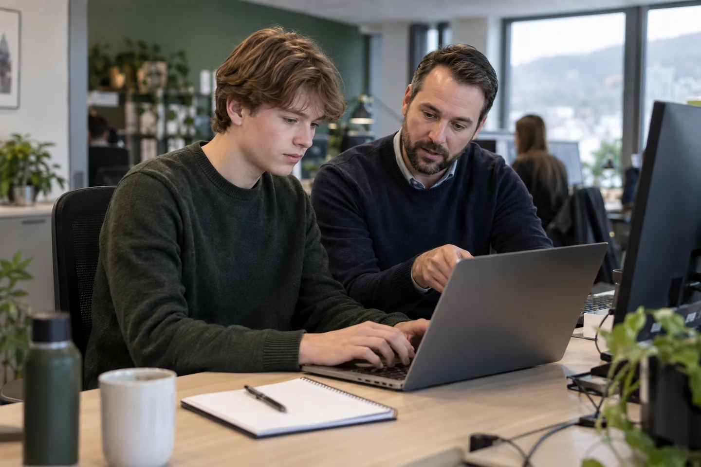 KI-generert illustrasjon av en praksiselev som arbeider på en bærbar PC med veiledning fra en kollega i en bedrift